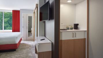A hotel room with a bed, a TV mounted on the wall, a small table with a remote control, a coffee maker, and a wardrobe.