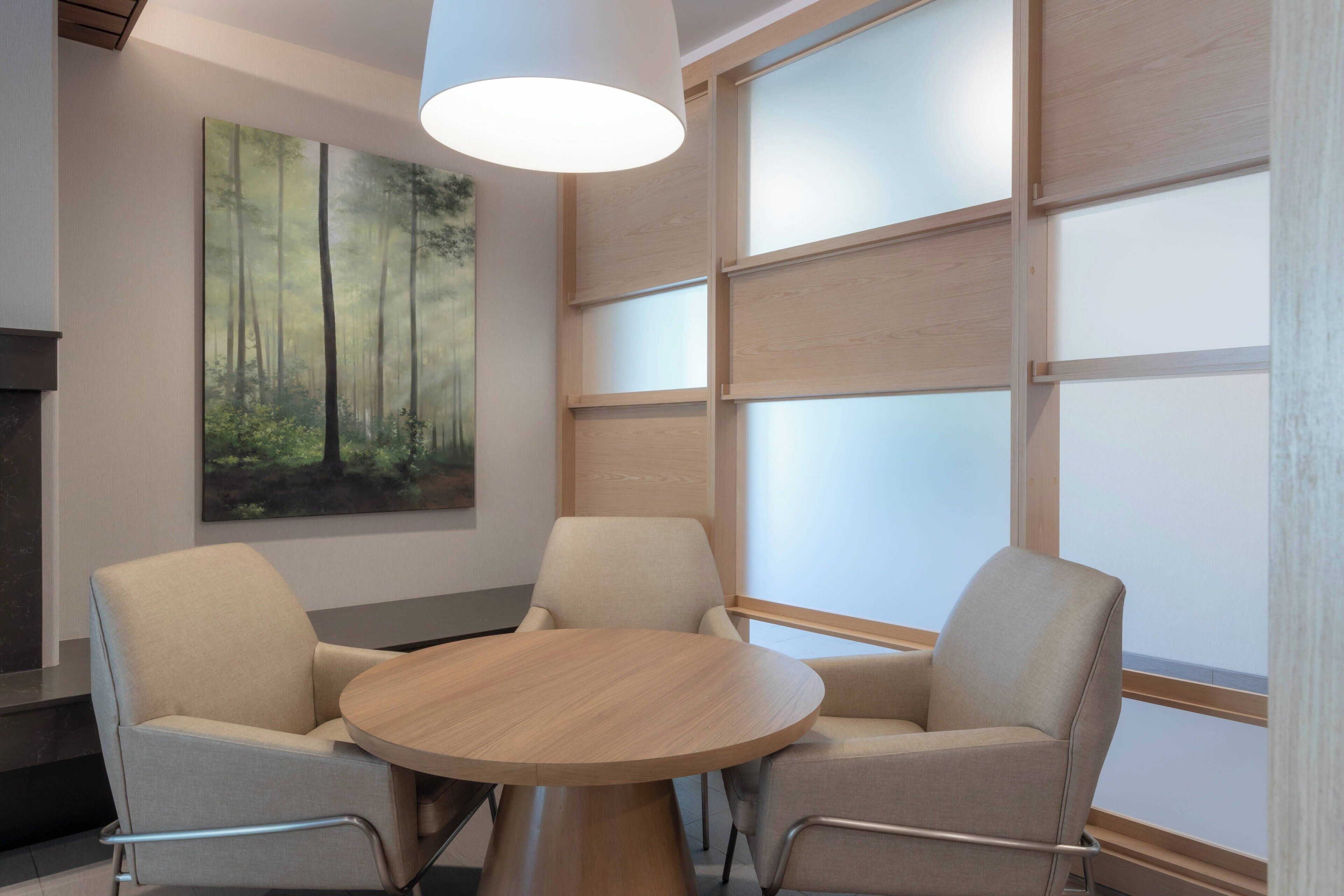 Ein moderner Konferenzraum mit einem runden Holztisch, zwei beigen Stühlen, einem großen Wandbild mit Waldmotiv und einem großen Fenster mit mattierten Glasscheiben.