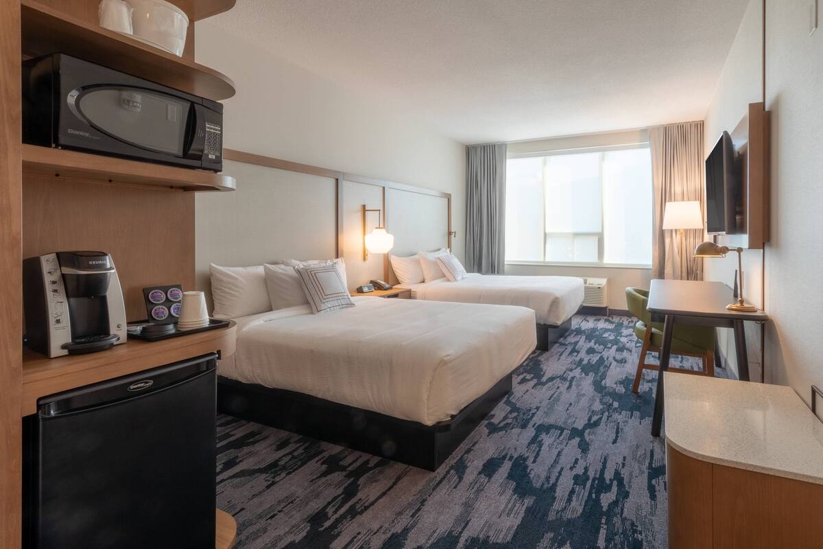 A hotel room with two beds, a microwave, a coffee maker, a refrigerator, a TV, and a window with curtains.