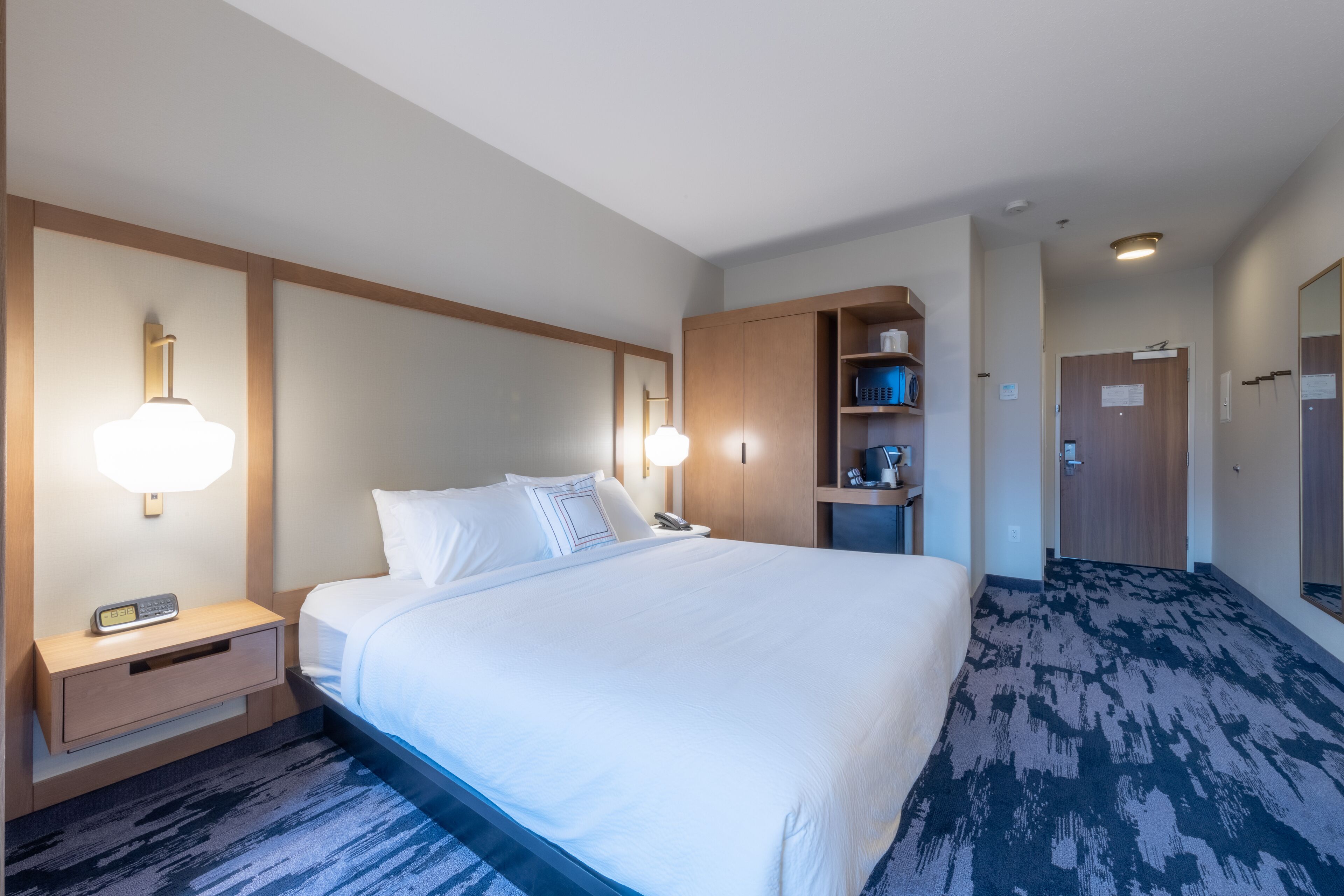 Hotelkamer met een groot bed, nachtkastjes, een lamp, een plank met toiletartikelen en een deur naar een andere kamer.