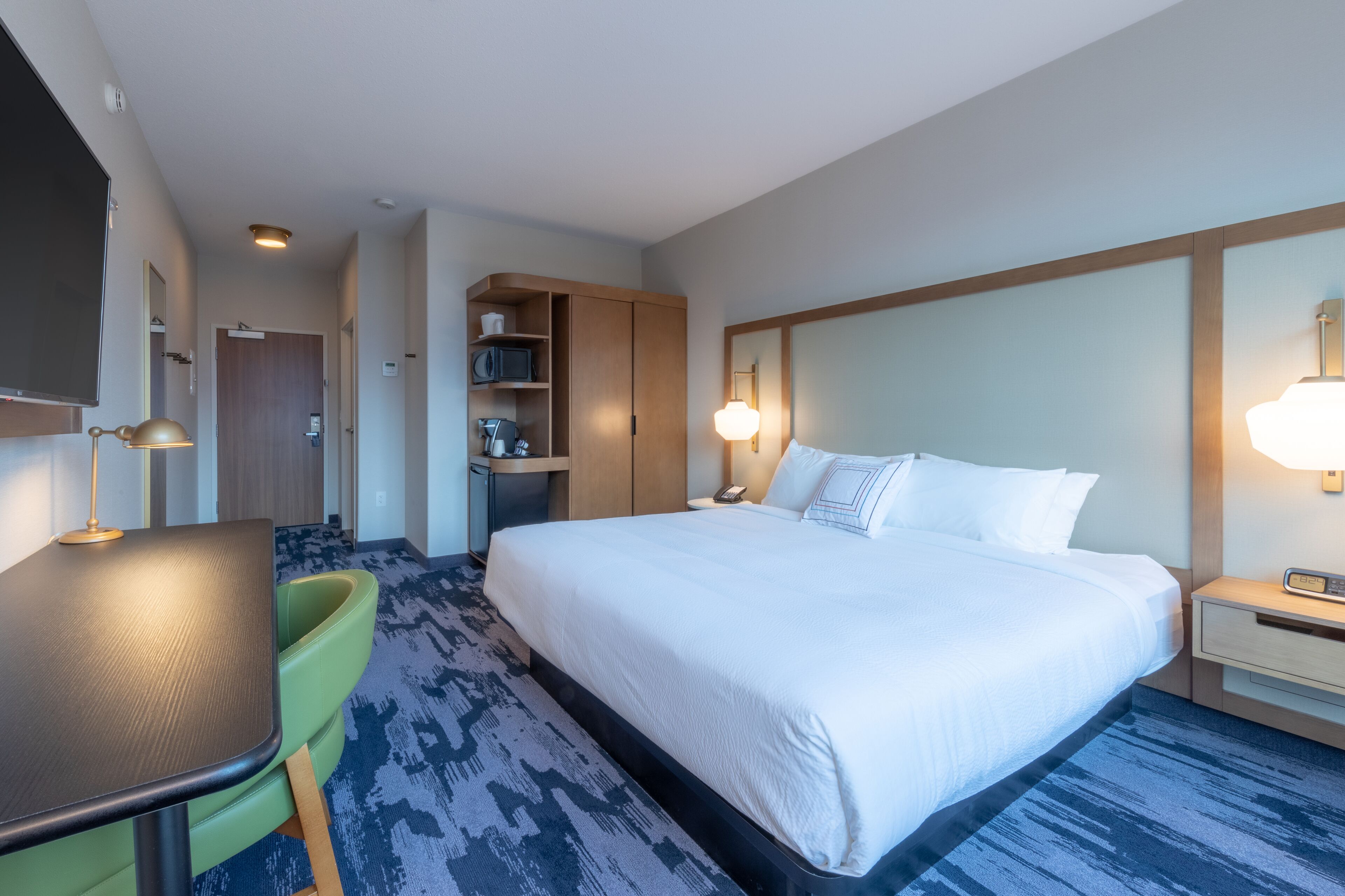 A hotel room with a large bed, a desk, a chair, a small kitchenette, and a bathroom.