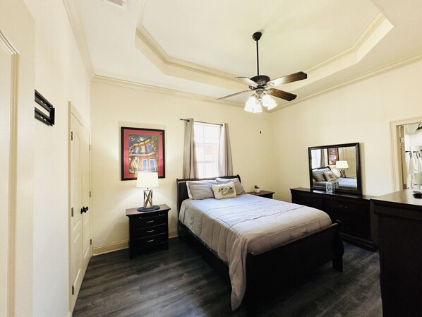 Une chambre à coucher comprenant un lit, des tables de chevet, un ventilateur de plafond, une fenêtre avec des rideaux et un miroir fixé au mur.