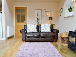 Ein gemütliches Wohnzimmer mit einem braunen Ledersofa, zwei gemusterten Kissen, einem violetten Shaggy-Teppich, einer Holztür, einem weißen Heizkörper, einem an der Wand befestigten Regal mit einer Pflanze, gerahmten Bildern und einem schwarzen Kaminofen.