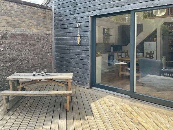 A wooden deck with a picnic table and chairs, a brick wall, and a glass door leading to an interior space with a kitchen and seating area.