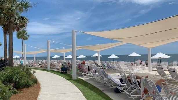 Un espace de détente aménagé au bord de la plage, avec plusieurs rangées de chaises longues à l’ombre de grands parasols, un chemin en béton et des palmiers.