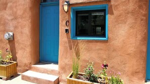 Un bâtiment recouvert d’enduit, avec une porte et une fenêtre bleues, un petit porche avec des marches et un jardinier avec des plantes.