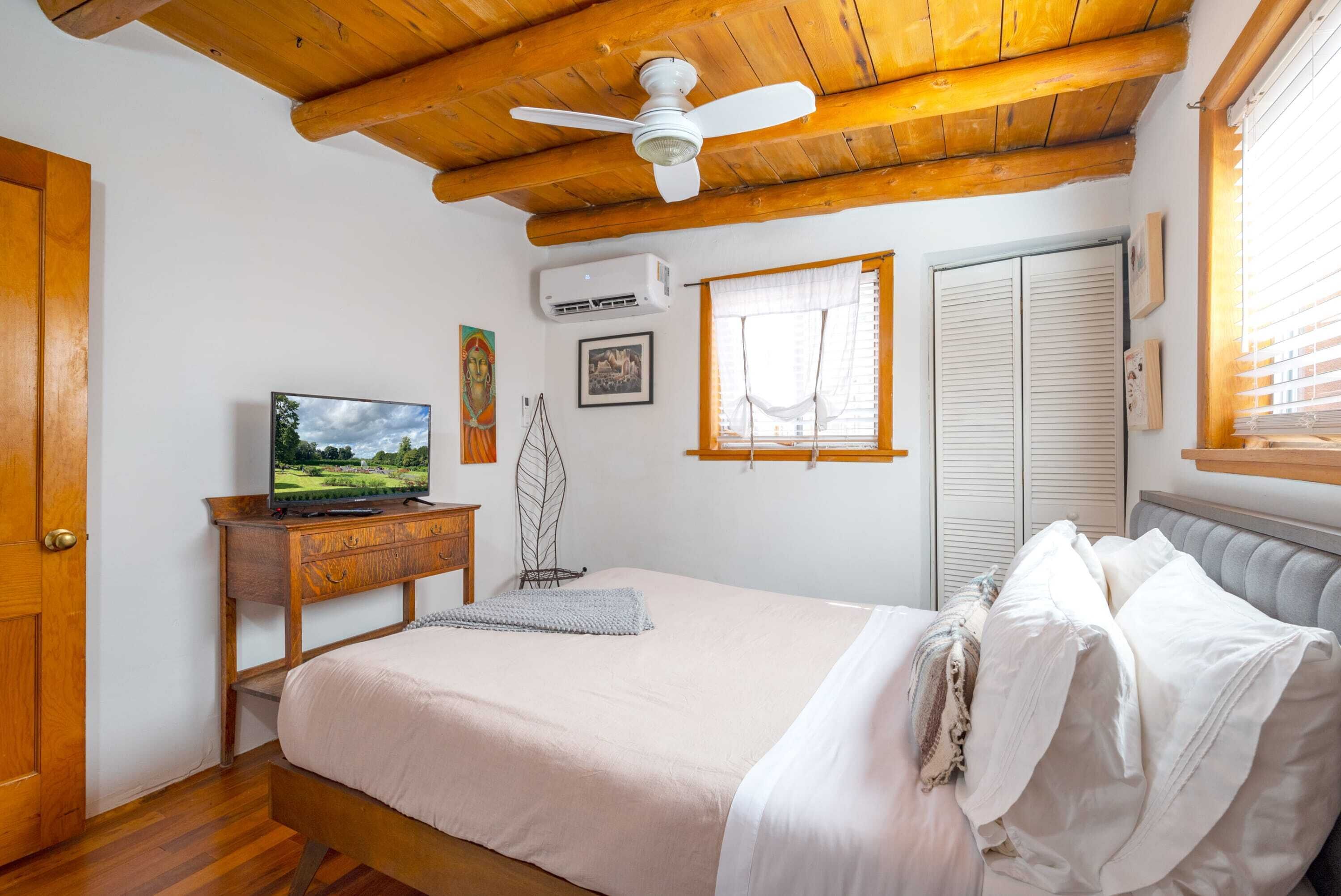 A bedroom with a bed, wooden ceiling, ceiling fan, TV, air conditioning unit, window with blinds, and a wardrobe.