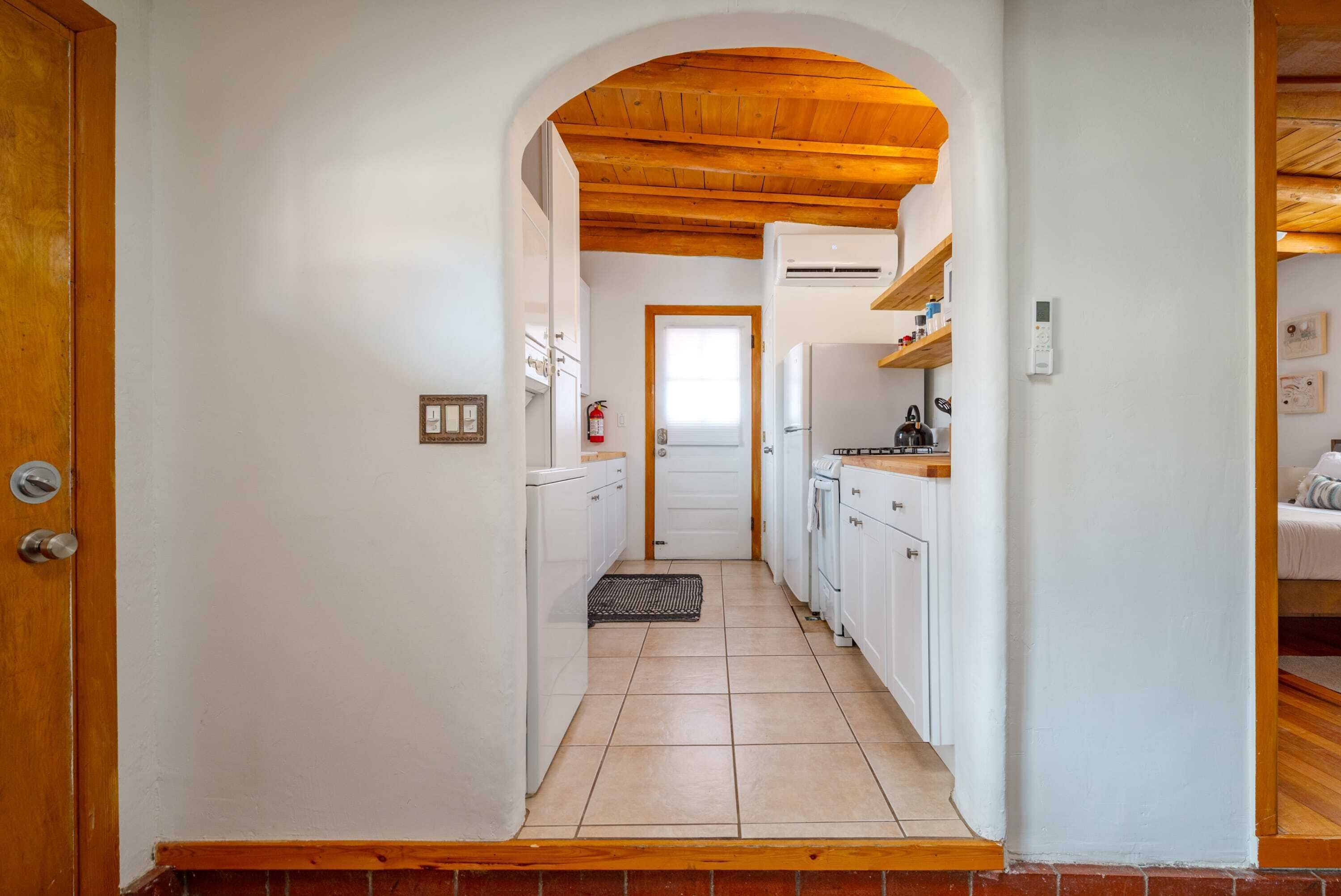 A narrow hallway leading to a kitchen with white cabinets, a wooden ceiling, and a door at the end.