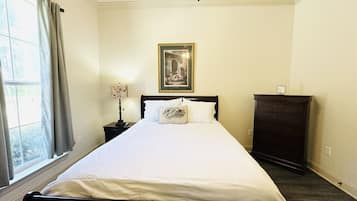 A neatly made bed in a room with a window, a framed picture on the wall, and a dresser.