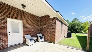Een bakstenen gebouw met een witte deur, twee rieten stoelen met kussens, en een betonnen terras.