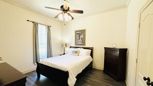 Een slaapkamer met een bed, een plafondventilator, een dressoir en een raam met gordijnen.