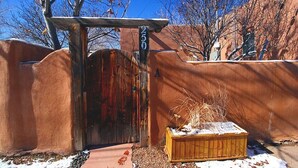 A rustic wooden gate with a metal number sign, a wooden planter with dried plants, and a clear blue sky.