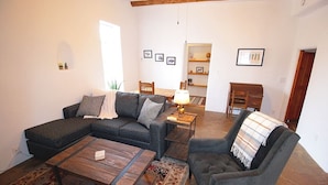 Sala de estar com sofá seccional, uma poltrona, uma mesa de centro e uma pequena mesa com abajur. Um tapete cobre o chão, e a sala possui paredes brancas com uma viga de madeira no teto.