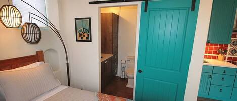 A bedroom with a bed, a door leading to a bathroom, and a kitchenette area.