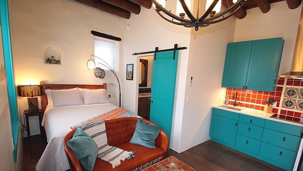 Un dormitorio con cama, sofá, una mesita y una zona de cocina con armarios color turquesa y salpicadero rojo.