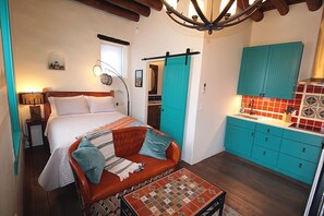 Een slaapkamer met een bed, een bank, een kleine tafel en een keukenhoek met blauwgroene kastjes en een rode achterwand.