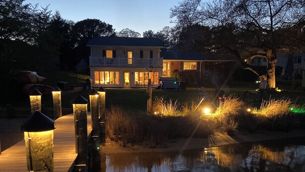 A wooden dock extending over a calm body of water, leading to a house with a balcony and large windows.