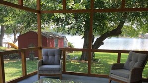 A covered patio area with wicker furniture and a glass table, overlooking a lake and a red building.
