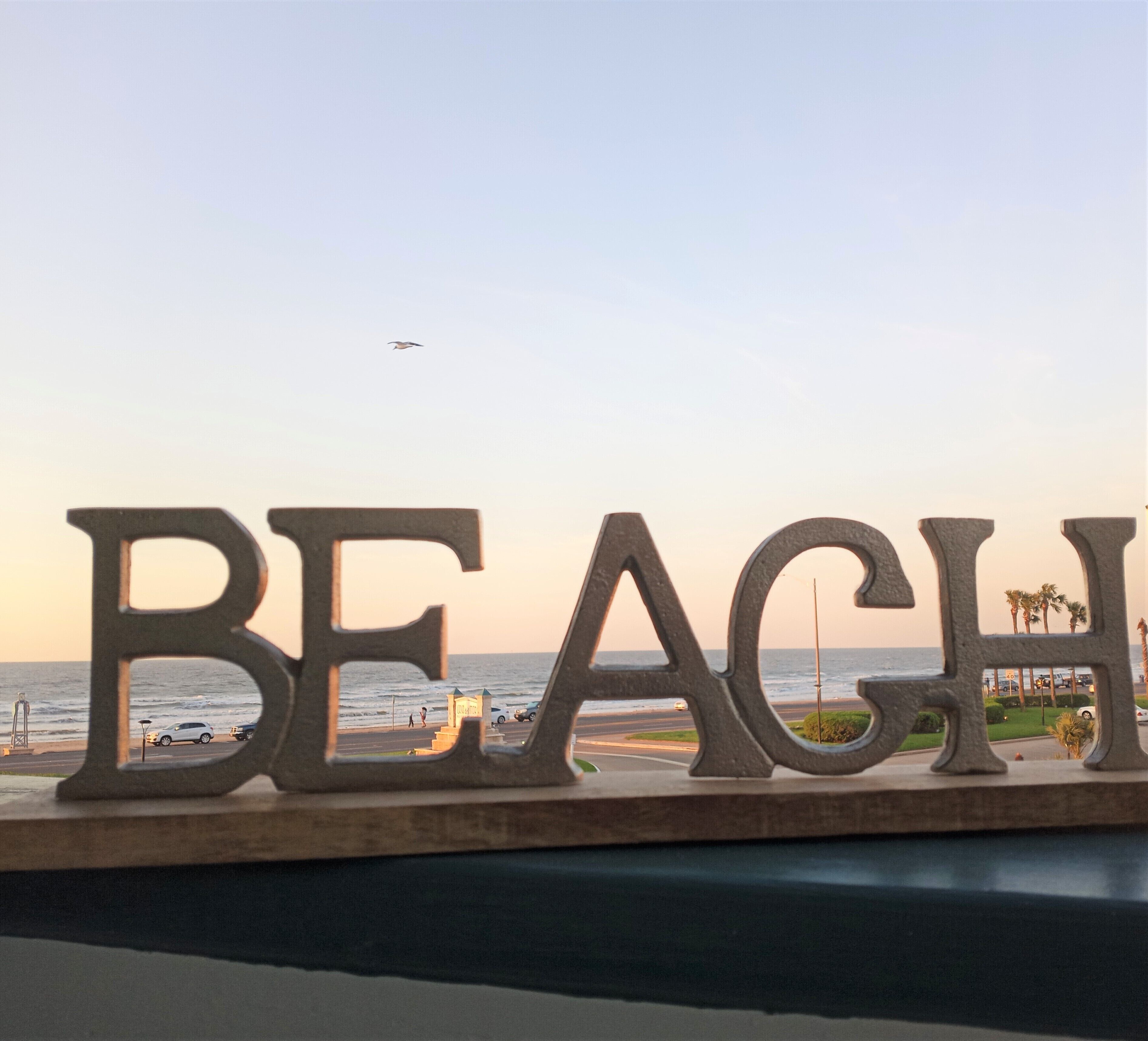 A sign with the word "BEACH" in large letters, overlooking a coastal area with a clear sky and a plane in the distance.
