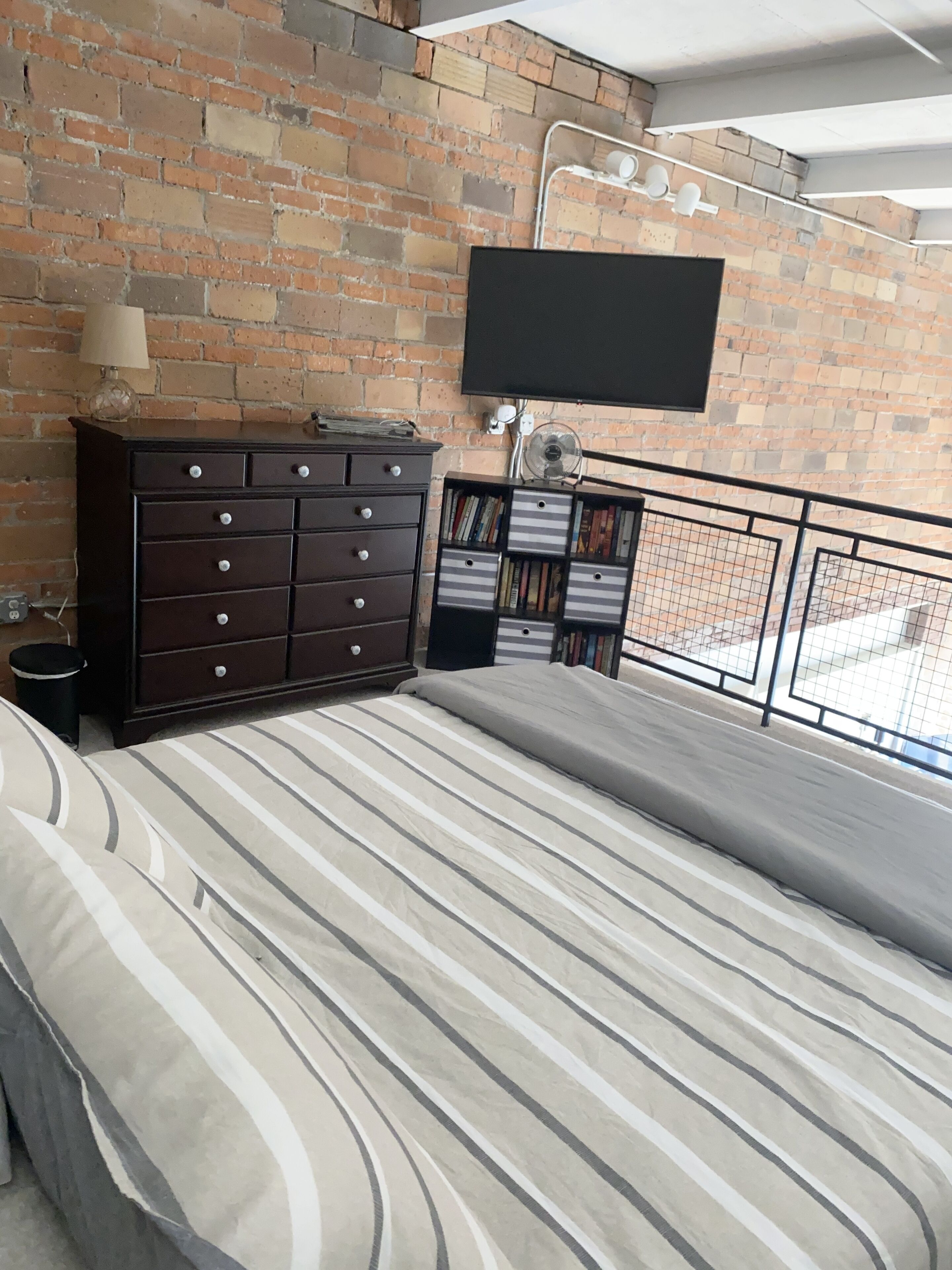 A bedroom with a brick wall, a flat-screen TV mounted above a dresser, a bookshelf, and a bed with striped bedding.