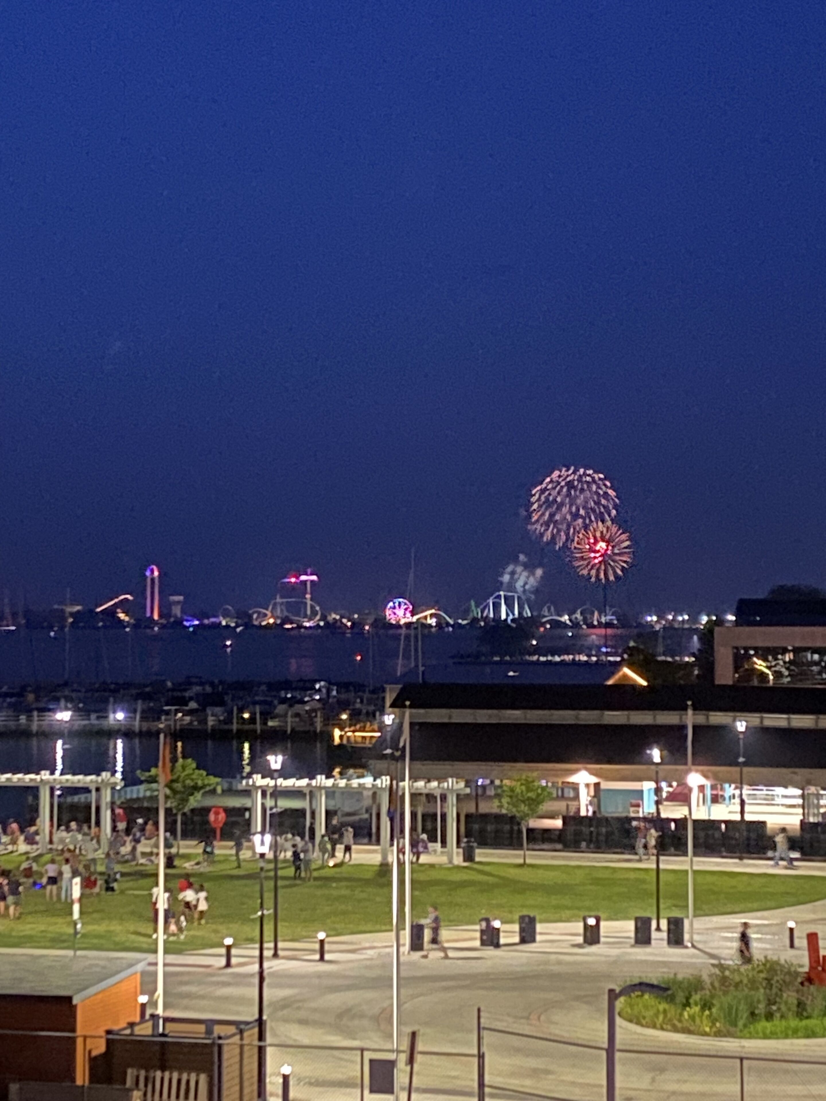 Vista nocturna de una zona costera con un espectáculo de fuegos artificiales, una noria y una multitud de gente.