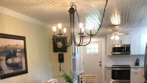 Un coin repas avec une table en bois dressée pour six personnes, entourée de chaises blanches. Un lustre est suspendu au plafond, et un tableau représentant une vache est accroché au mur.