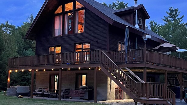 A two-story wooden house with a covered porch and a staircase leading to the upper level. - Warm Secluded Adirondack Chalet with Game Room and Bar/Lounge (Warrensburg)