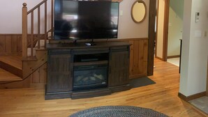 A living room with a wooden staircase, a flat-screen TV on a wooden entertainment unit, a round mirror on the wall, and a patterned rug on the floor. - Warm Secluded Adirondack Chalet with Game Room and Bar/Lounge (Warrensburg)