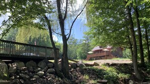 A wooden bridge over a rocky stream, leading to a large wooden house with a balcony. - Warm Secluded Adirondack Chalet with Game Room and Bar/Lounge (Warrensburg)