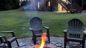 A stone fire pit with chairs, a wooden deck, and a house with a balcony and string lights. - Warm Secluded Adirondack Chalet with Game Room and Bar/Lounge (Warrensburg)