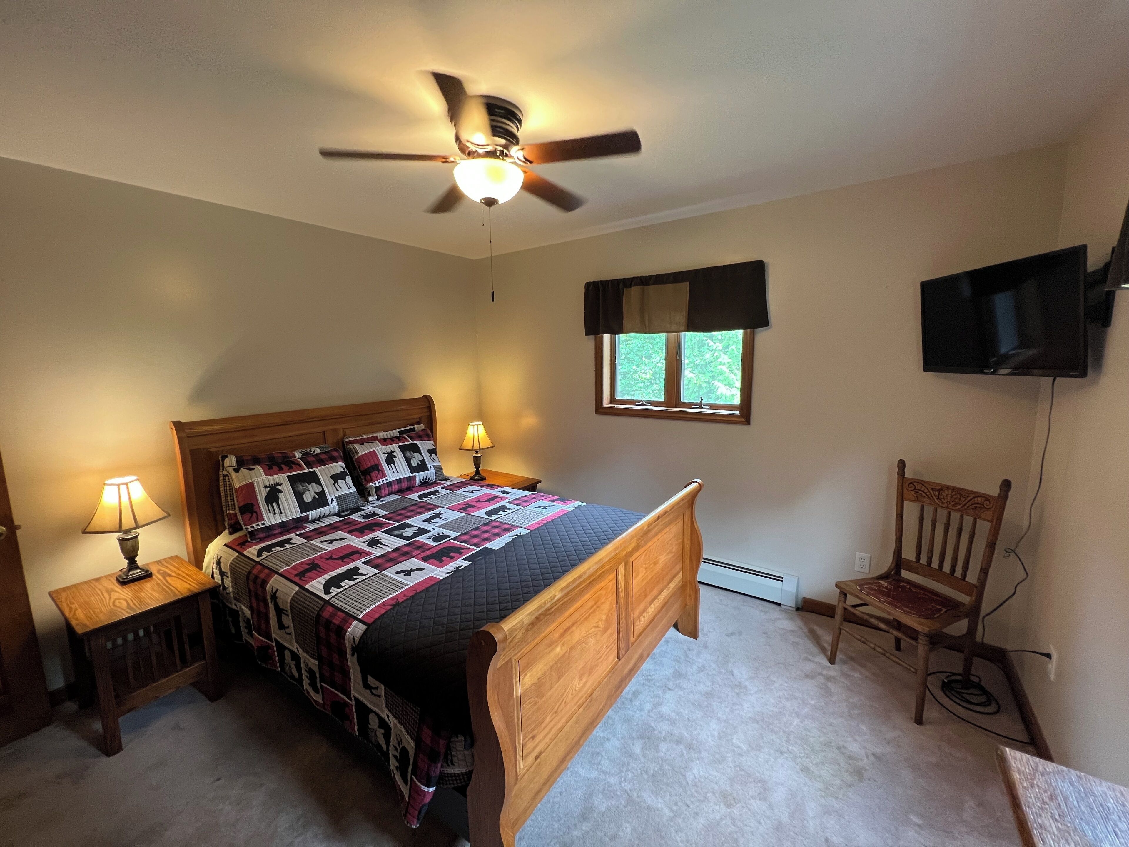A bedroom with a bed, a ceiling fan, a nightstand with a lamp, a window with a view, and a TV mounted on the wall.