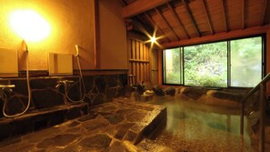 A hot spring bath with stone flooring, wooden benches, and a view of greenery through a large window. - Usuki Yunosato (Usuki)