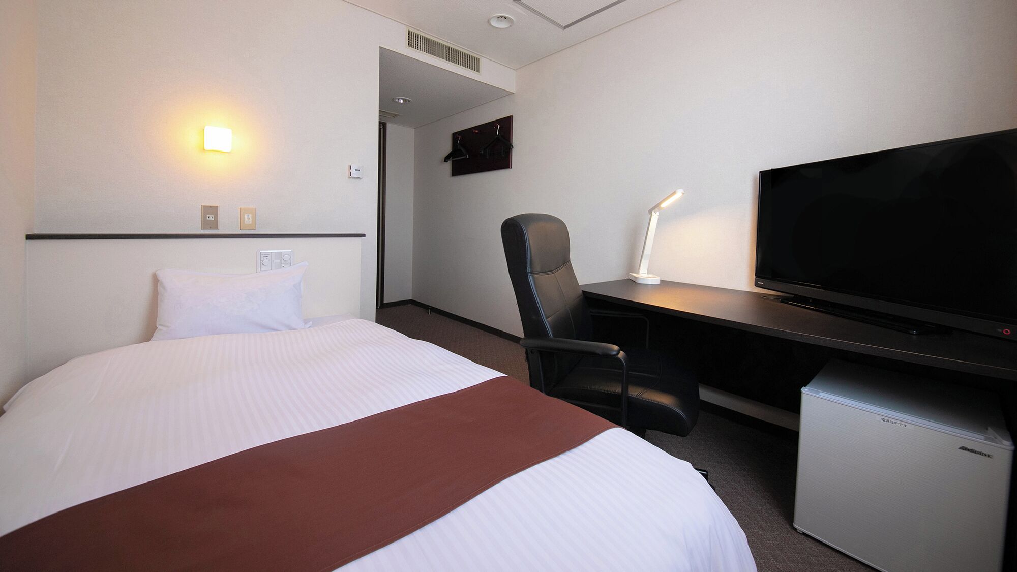 A hotel room with a bed, a desk with a television, a chair, and a small refrigerator.