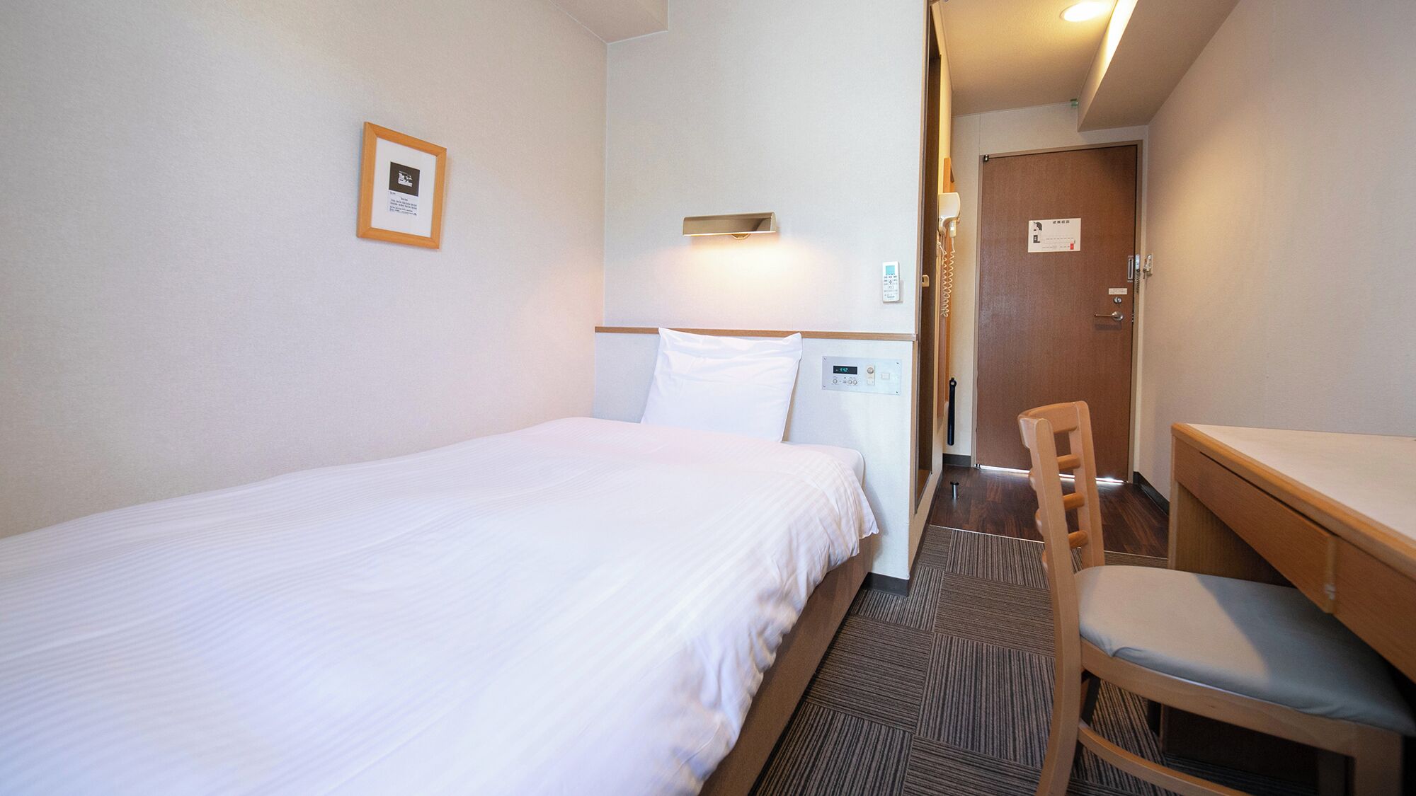 A hotel room with a bed, a desk, a chair, a bedside table, a nightstand, a wall-mounted lamp, and a door.