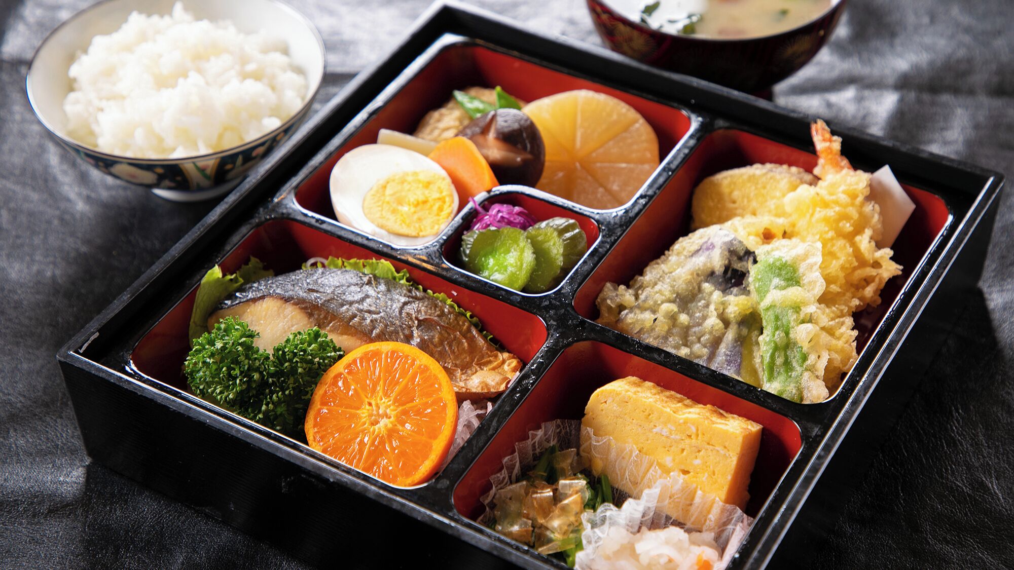 A bento box with various food items including fish, egg, vegetables, and rice.