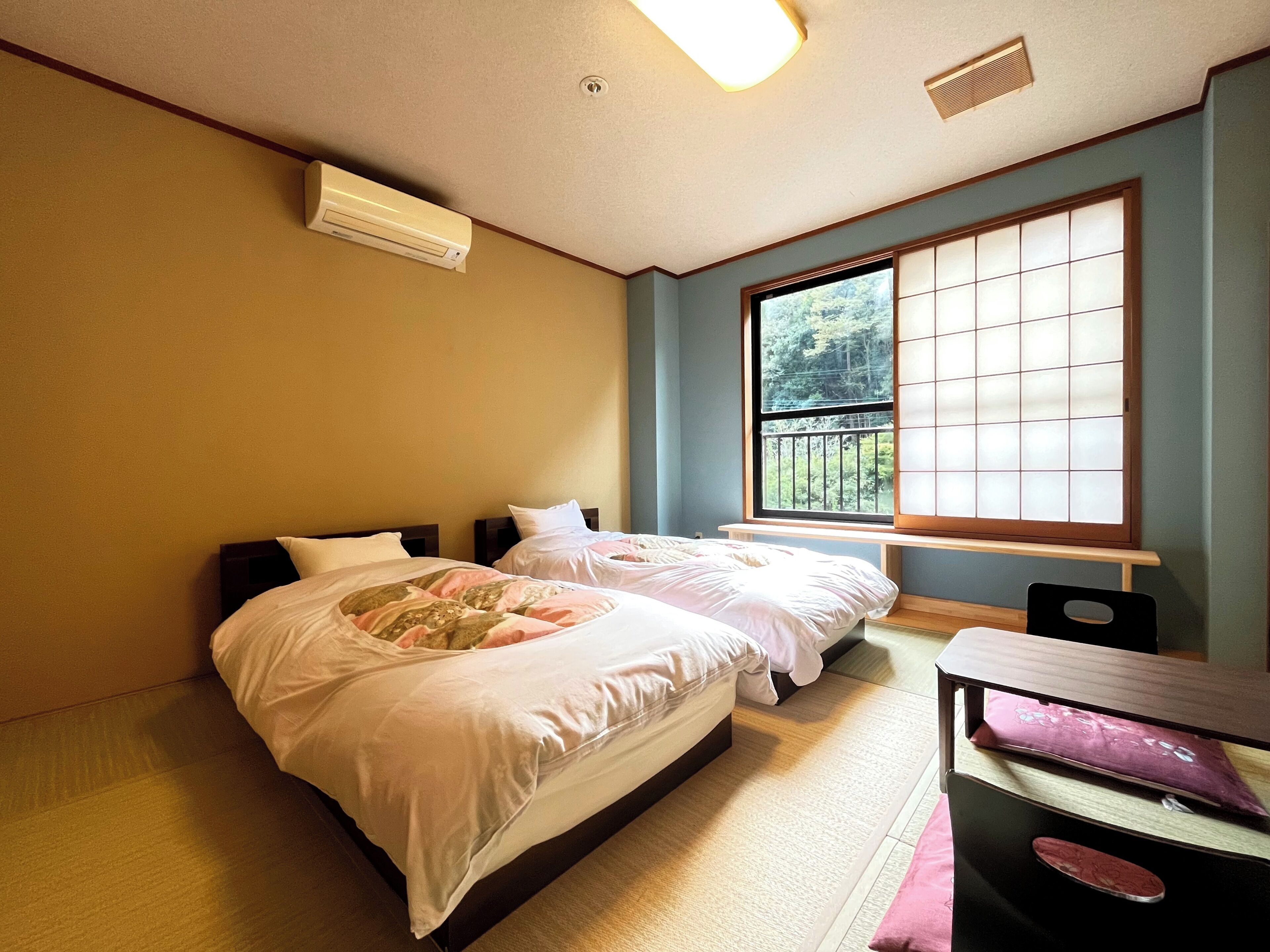 A bedroom with two beds, a window with a view of greenery, an air conditioning unit, and a small table with a chair.