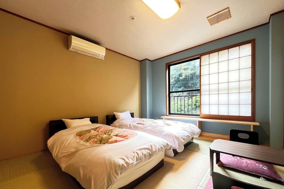 Une chambre avec deux lits, une fenêtre donnant sur un espace verdoyant, un appareil de climatisation et une petite table avec une chaise.