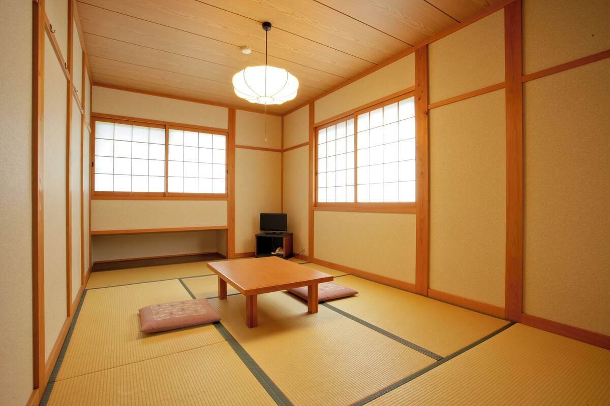 Une pièce de style japonais traditionnel, avec un sol recouvert de tatamis, une table basse, des coussins, des portes coulissantes et un luminaire suspendu au plafond.