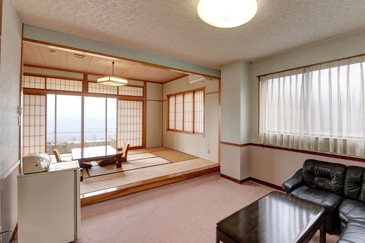 A traditional Japanese room with tatami flooring, a tatami mat room, a black leather sofa, a small table, and a view of the sea.