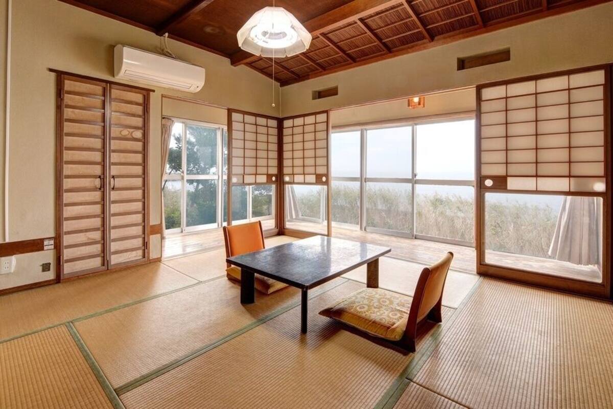 Una habitación con suelo de tatami, con una mesa y sillas, ventanas amplias y un ventilador de techo.