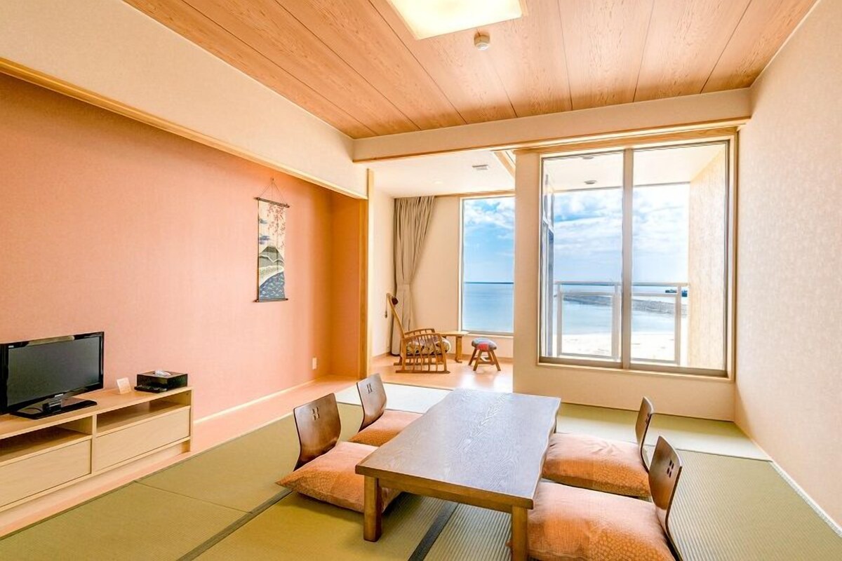 A room with tatami mats, a wooden table, a television, and a large window offering a view of the sea.