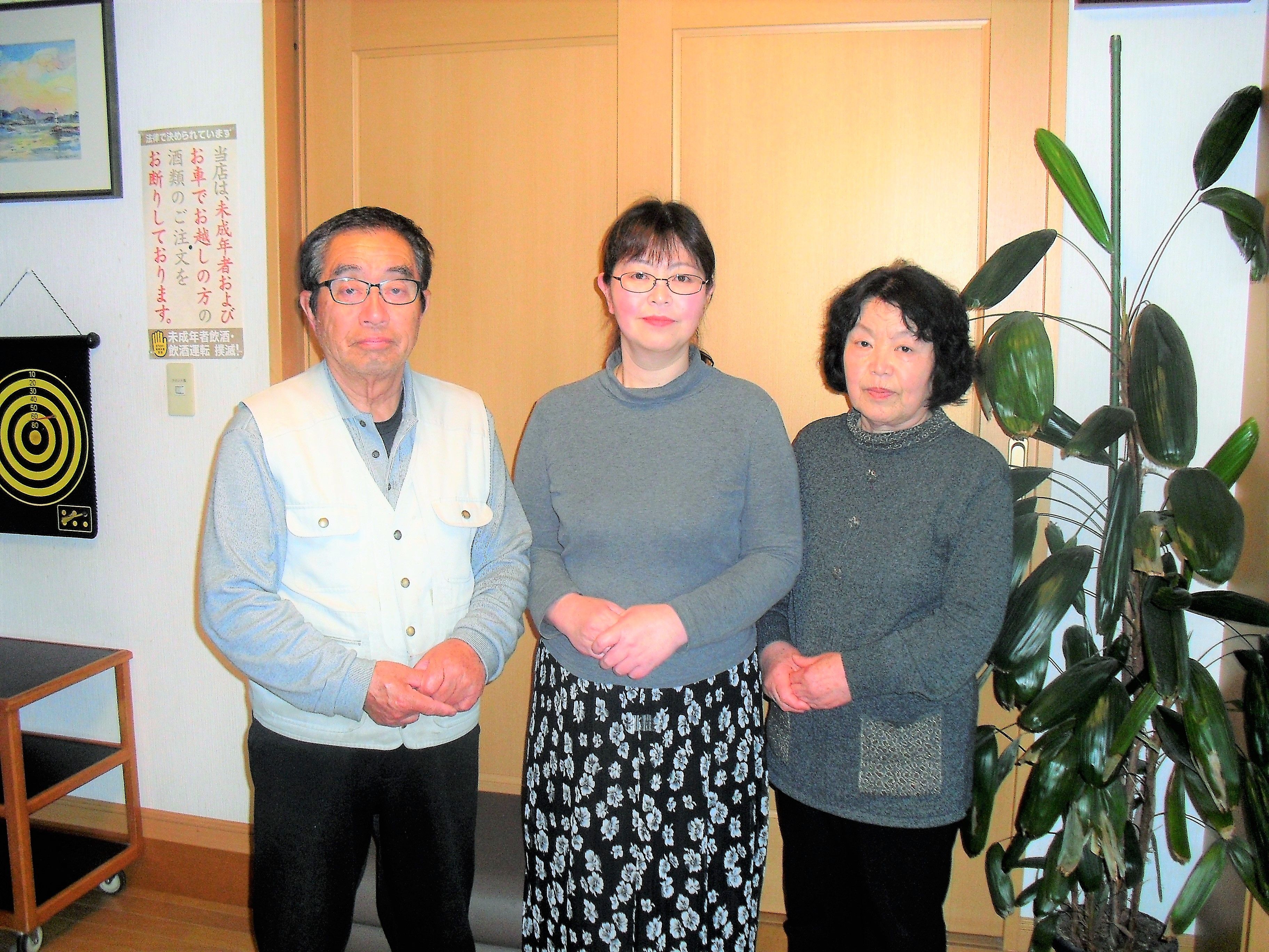 Three individuals standing indoors with a dartboard and a plant in the background.