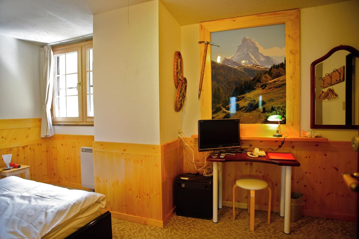 A room with a bed, a desk with a computer, a chair, a window with curtains, and a painting of a mountain landscape.