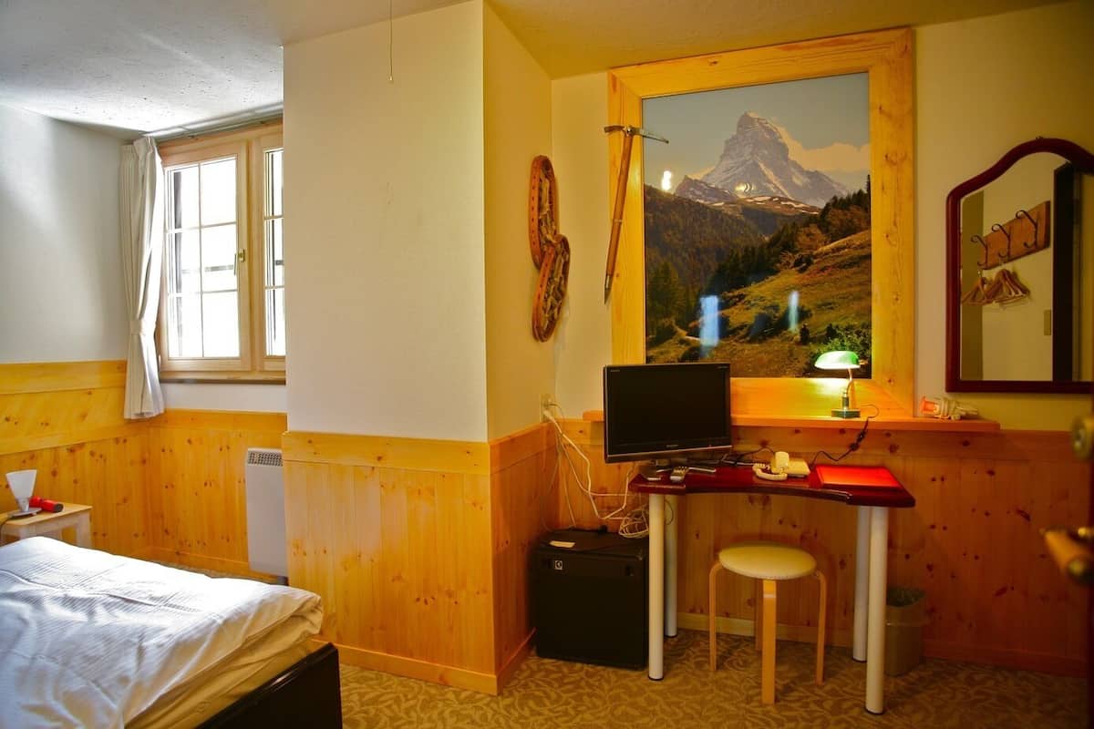 A room with a bed, a desk with a computer, a chair, a window with curtains, a painting of a mountain, and a mirror.