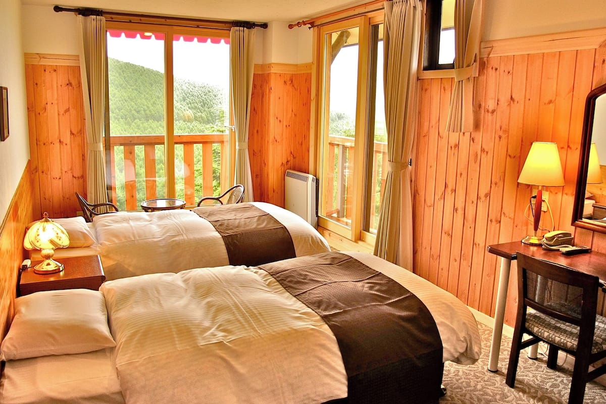A room with two beds, a desk with a lamp, a chair, and a window with curtains.