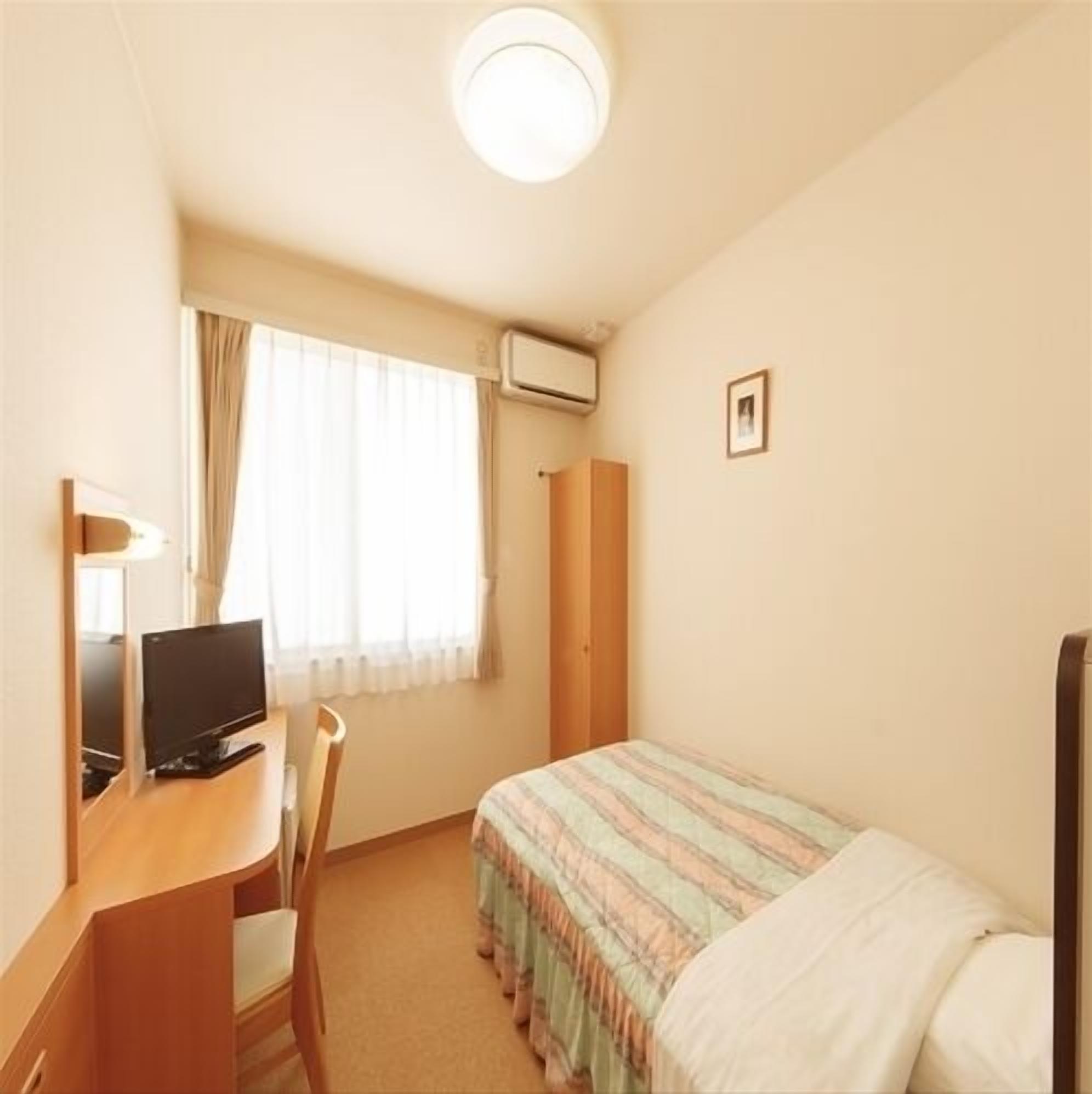 Pieni hotellihuone, jossa on sänky, työpöytä tietokoneella, tuoli, ikkuna verhoilla ja taulu seinällä.