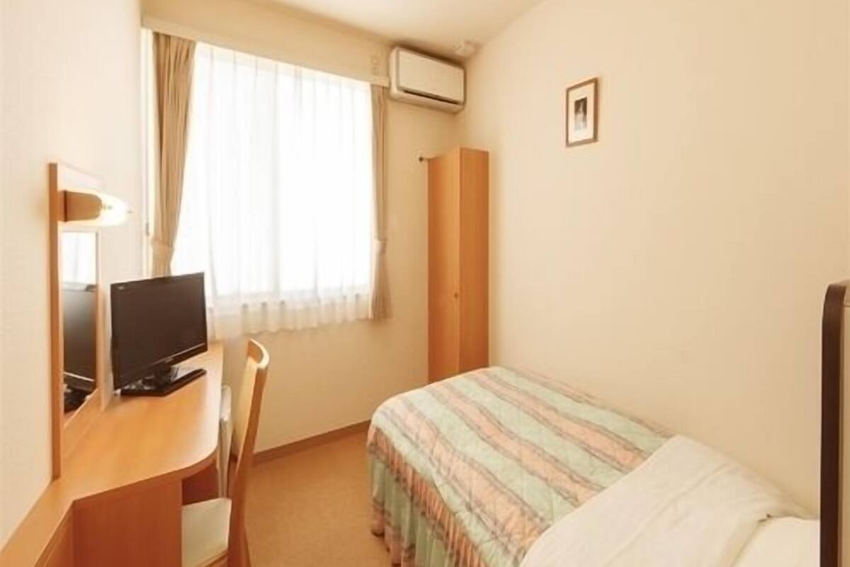 Ein kleines Hotelzimmer mit Bett, Schreibtisch mit Computer, Stuhl, Fenster mit Vorhängen und einem Bild an der Wand.