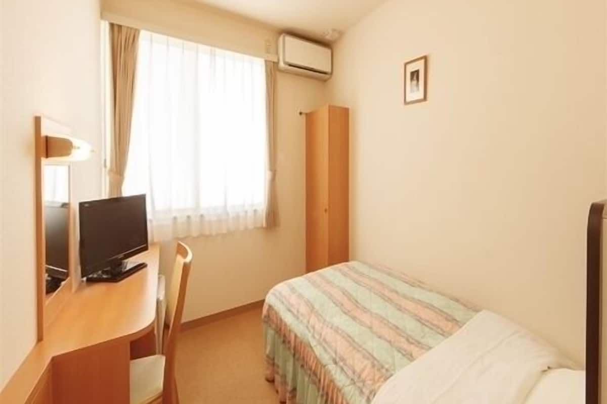Pieni hotellihuone, jossa on sänky, työpöytä tietokoneella, tuoli, ikkuna verhoilla ja taulu seinällä.