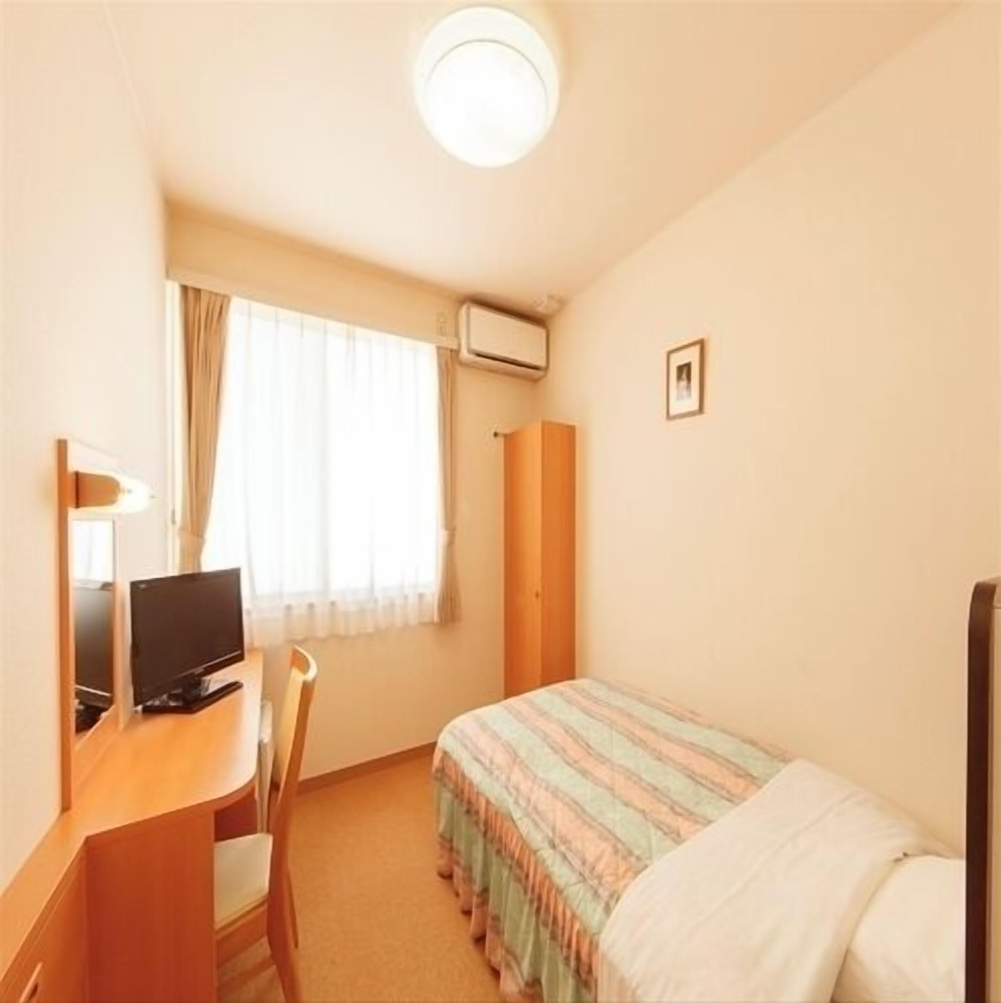 A small hotel room with a single bed, a desk with a computer, a chair, a window with curtains, and a wall-mounted air conditioner.