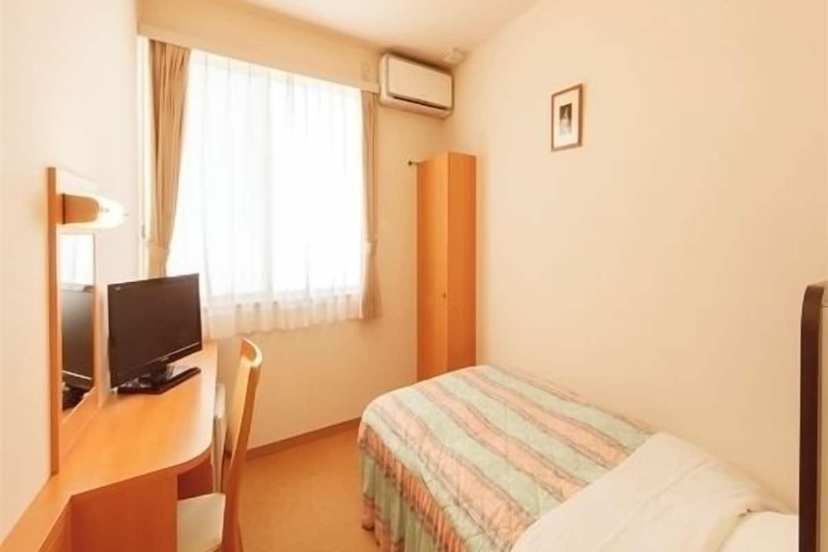 Ein kleines Hotelzimmer mit einem Einzelbett, einem Schreibtisch mit Computer, einem Stuhl, einem Fenster mit Vorhängen und einer Wand-Klimaanlage.